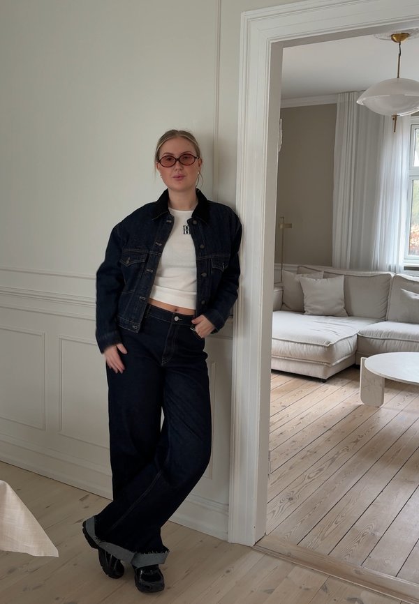Temno moder jeans jakna s črnim ovratnikom, bela kratka majica in temno široke kavbojke z neobdelanimi robovi. Krogli roza sončna očala dopolnjujejo obleko.