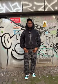 Persoană tânără îmbrăcată într-un hanorac negru, pantaloni cu model și adidași, stând cu mâinile în buzunare, în fața unui perete acoperit cu graffiti și semne de avertizare.