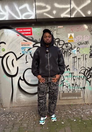 Joven con sudadera negra, pantalones estampados y zapatillas de deporte, de pie con las manos en los bolsillos frente a una pared cubierta de graffiti con señales de advertencia.