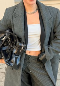 Γυναίκα που φοράει γκρι σακάκι, παντελόνι με ρίγες, λευκό crop top, μαύρο δερμάτινο γάντι και κολιέ, κρατώντας μια μεγάλη μαύρη δερμάτινη τσάντα.