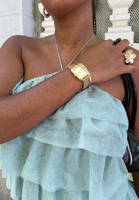 Gestufte, hellgrüne Rüschenshirt; goldene rechteckige Uhr mit Mesh-Armband; Blumenring; strukturierte schwarze Handtasche; Hautton ist mittel.