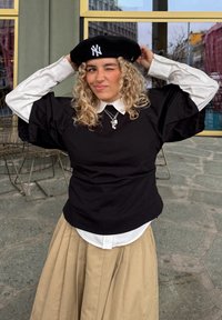 Sort topp med korte ermer over en hvit skjorte, kombinert med et beige plissert skjørt og en svart beret med hvit logo. Teksturert, avslappet stil.