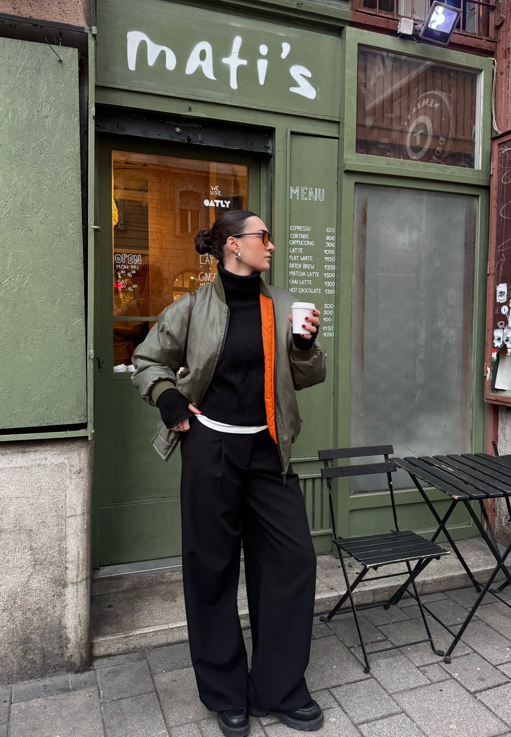 Femme en col roulé noir, veste bomber olive et lunettes de soleil tenant une tasse de café près d'une porte verte de café avec un menu et une table pliante noire avec des chaises.