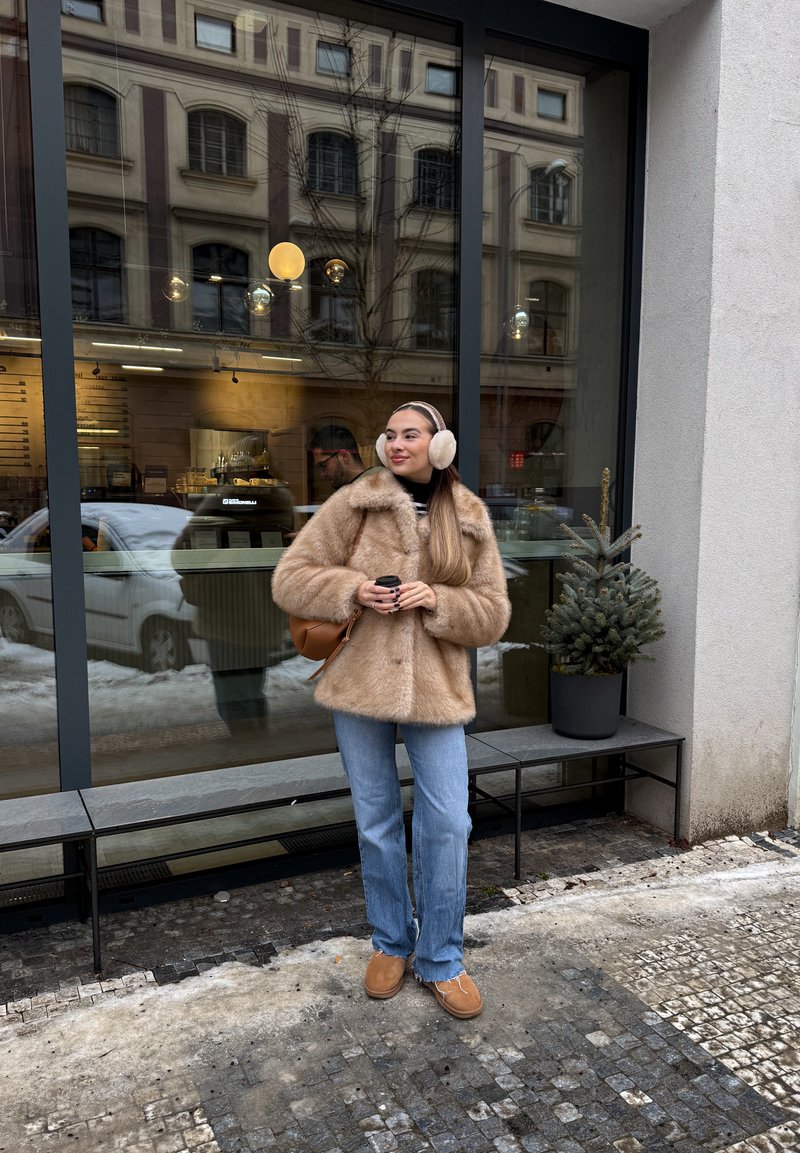 Young woman in beige fur coat and earmuffs holding coffee cup, standing on cobblestone street outside cafe with large window and small potted pine tree.
