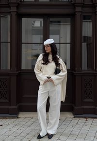 Biały, oversize sweter z szerokimi rękawami, kremowe spodnie typu wide-leg, czarny pasek, czarna kopertówka i biała beretka, na tle ciemnobrązowym.