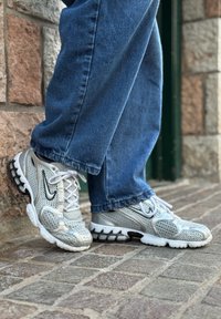 Sølvfargede treningssko i mesh-materiale, med teksturert design, synlige lisser og svarte detaljer, kombinert med blå, løstsittende jeans.