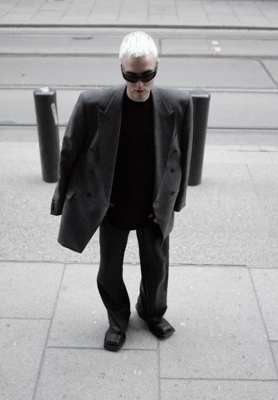 Persona con cabello corto rubio platino que lleva una blazer gris oversize, pantalones negros, camisa negra, zapatos de punta cuadrada y gafas de sol oscuras, de pie sobre la acera.