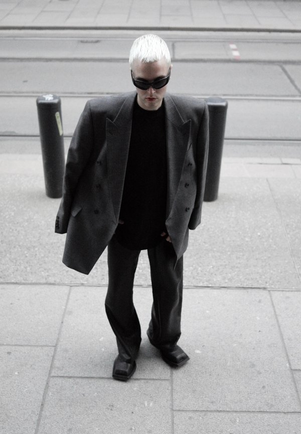 Persona con capelli corti biondo platino che indossa un blazer grigio oversize, pantaloni neri, camicia nera, scarpe con punta quadrata e occhiali da sole scuri, in piedi sul marciapiede.