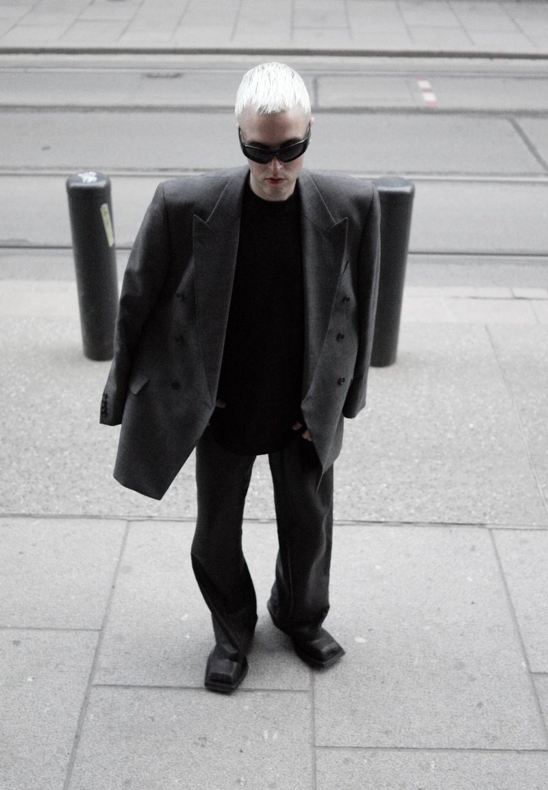 Personne aux cheveux courts blond platine portant un blazer gris surdimensionné, un pantalon noir, une chemise noire, des chaussures à bout carré et des lunettes de soleil foncées, debout sur le trottoir.