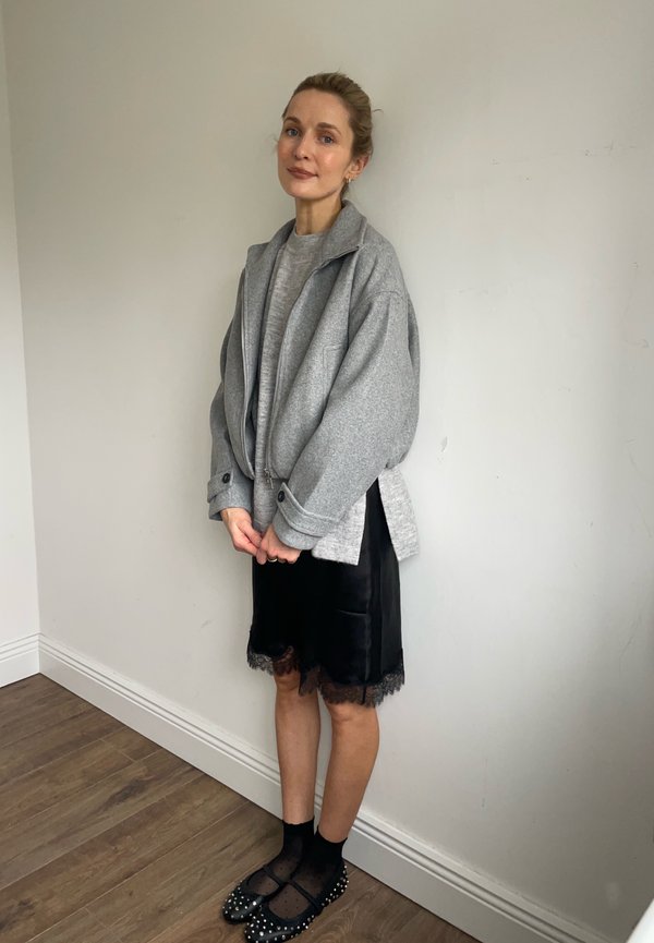 Naine seisab siseruumides tavalise seina taustal, kandes halli liibuva lukuga jopet, musta pitsist ääristusega seelikut, musti sokke ja naelutatud musti madalaid kingi.