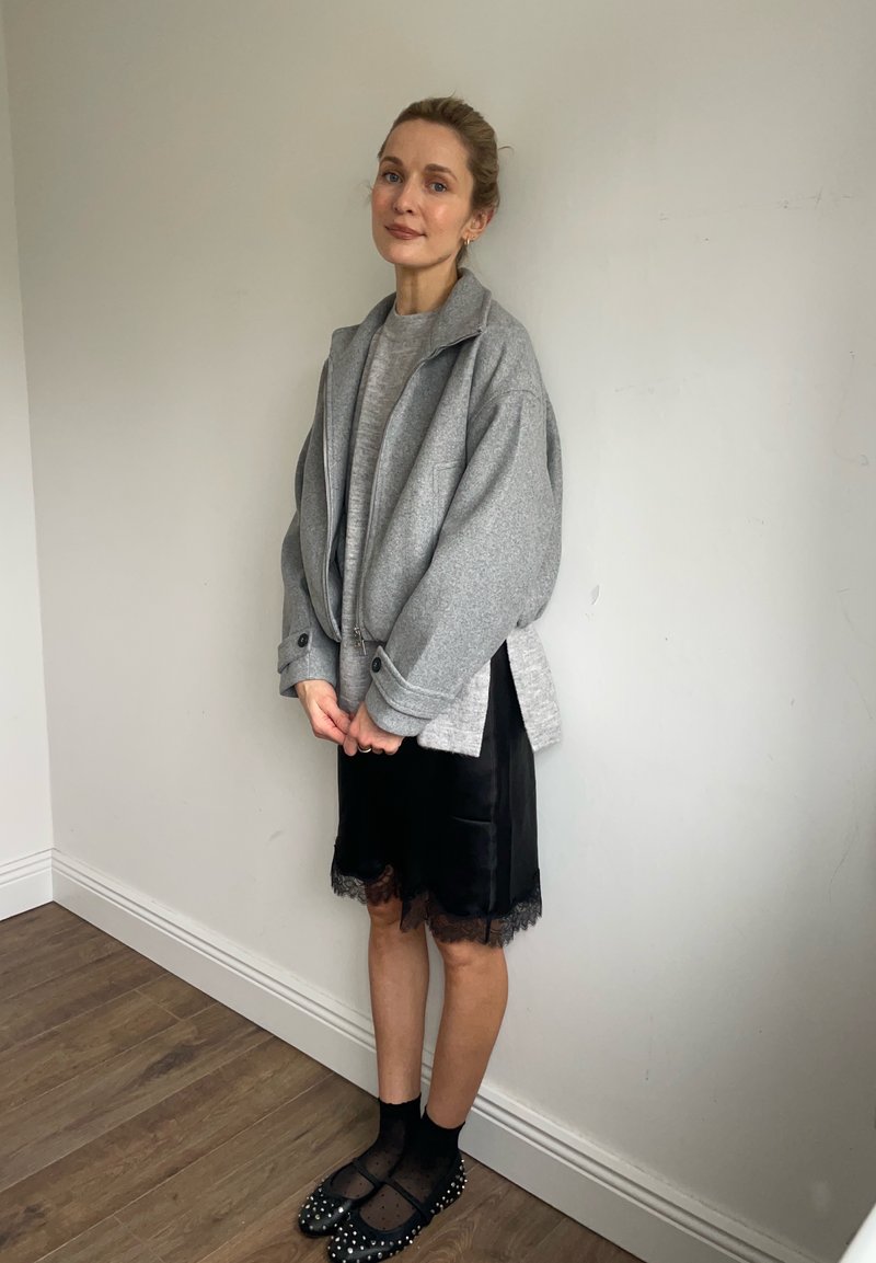 Une femme se tient à l'intérieur, contre un mur uni, vêtue d'une veste grise surdimensionnée à fermeture éclair, d'une jupe noire à bordure en dentelle, de chaussettes noires et de ballerines noires à clous.