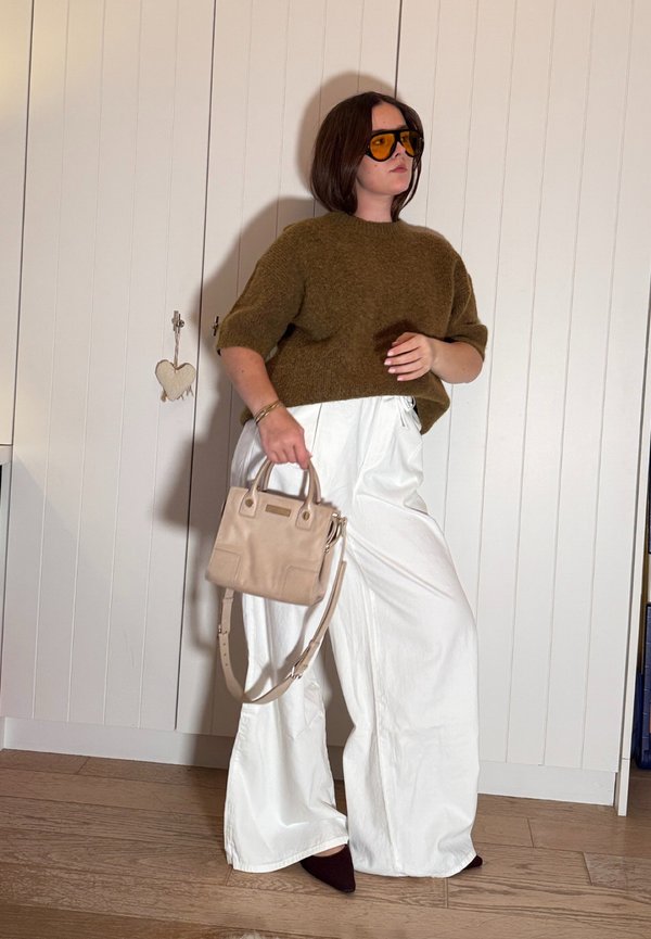 Femme portant un pull marron oversize, un pantalon blanc à jambes larges, des chaussures sombres à bouts pointus, des lunettes de soleil ambrées, tenant un petit sac à main beige à l'intérieur.