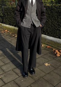 Trench coat preto sobre um colete cinza, camisa branca e gravata. Calças pretas soltas e sapatos pretos. Superfície pavimentada com folhas de outono.