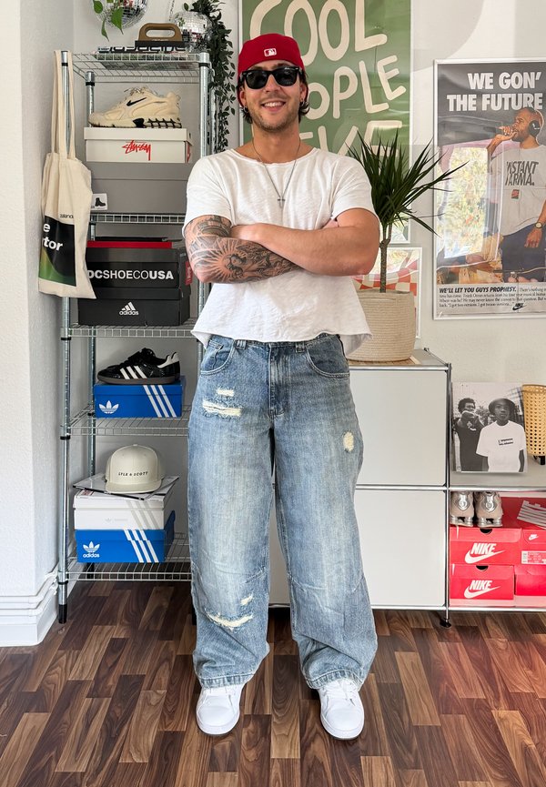 Hombre con gorra roja, gafas de sol, camiseta blanca y vaqueros rotos, de pie sonriendo con los brazos cruzados en una habitación con cajas de zapatos y carteles.