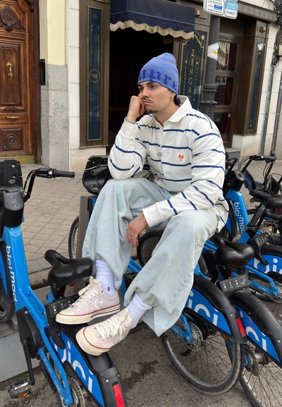 Joven con gorro azul, camisa blanca a rayas y pantalones claros sentado en bicicletas de alquiler compartidas azules en la acera de la ciudad, con expresión pensativa.