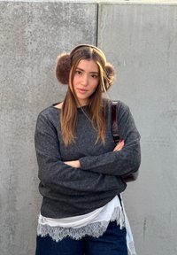 Jeune femme portant des cache-oreilles marron et un pull gris foncé, debout les bras croisés devant un mur texturé gris.