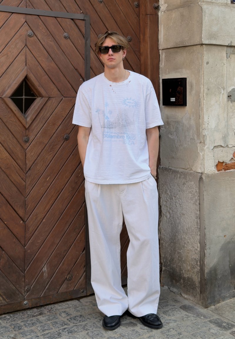 Jeune homme portant des lunettes de soleil, un t-shirt blanc ample et un pantalon, debout devant une grande porte en bois et un mur en pierre.