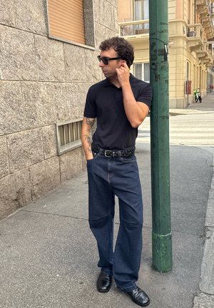 Hombre con tatuajes que lleva gafas de sol negras, polo negro y pantalones anchos, de pie en la acera junto a un poste verde en un entorno urbano.