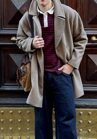 Personne portant un manteau beige, une chemise rayée rouge et noire, un pantalon bleu foncé, tenant un sac marron, se tenant contre une porte en bois sombre ornée de clous en laiton.