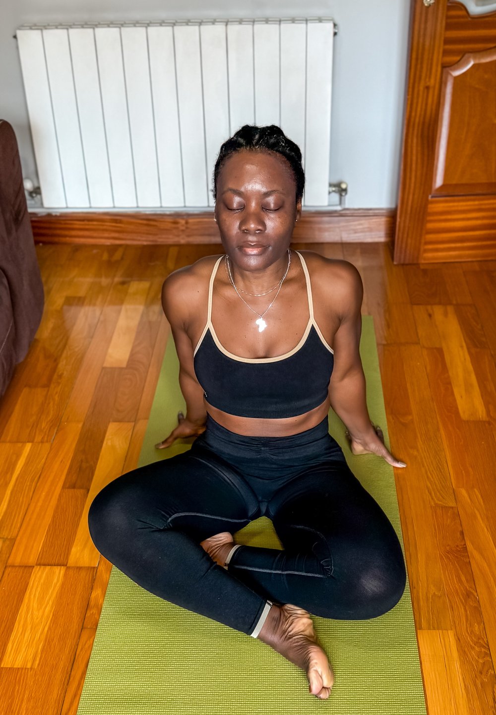 Femme vêtue de vêtements de sport noirs, assise en tailleur sur un tapis de yoga vert à l'intérieur, les yeux fermés, les mains posées au sol derrière elle.