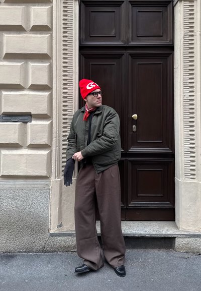 Hombre con gorro rojo, gafas, chaqueta verde, bandana roja y pantalones anchos marrones está de pie ajustándose un guante negro junto a una puerta de madera oscura.