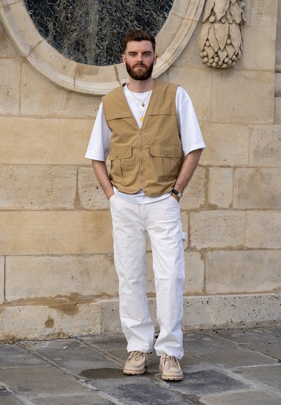 Chaleco beige sobre una camiseta blanca, pantalones cargo blancos y zapatillas beige. Cabello corto y barba. De pie frente a un fondo de pared de piedra.