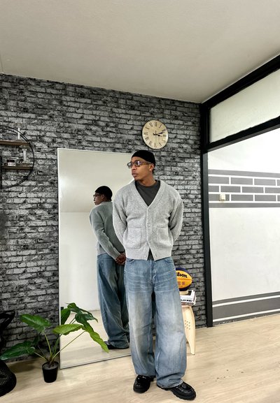 Cárdigan gris sobre una camisa oscura, combinado con jeans claros y sueltos y zapatos negros. La habitación cuenta con un gran espejo y una planta en una maceta.