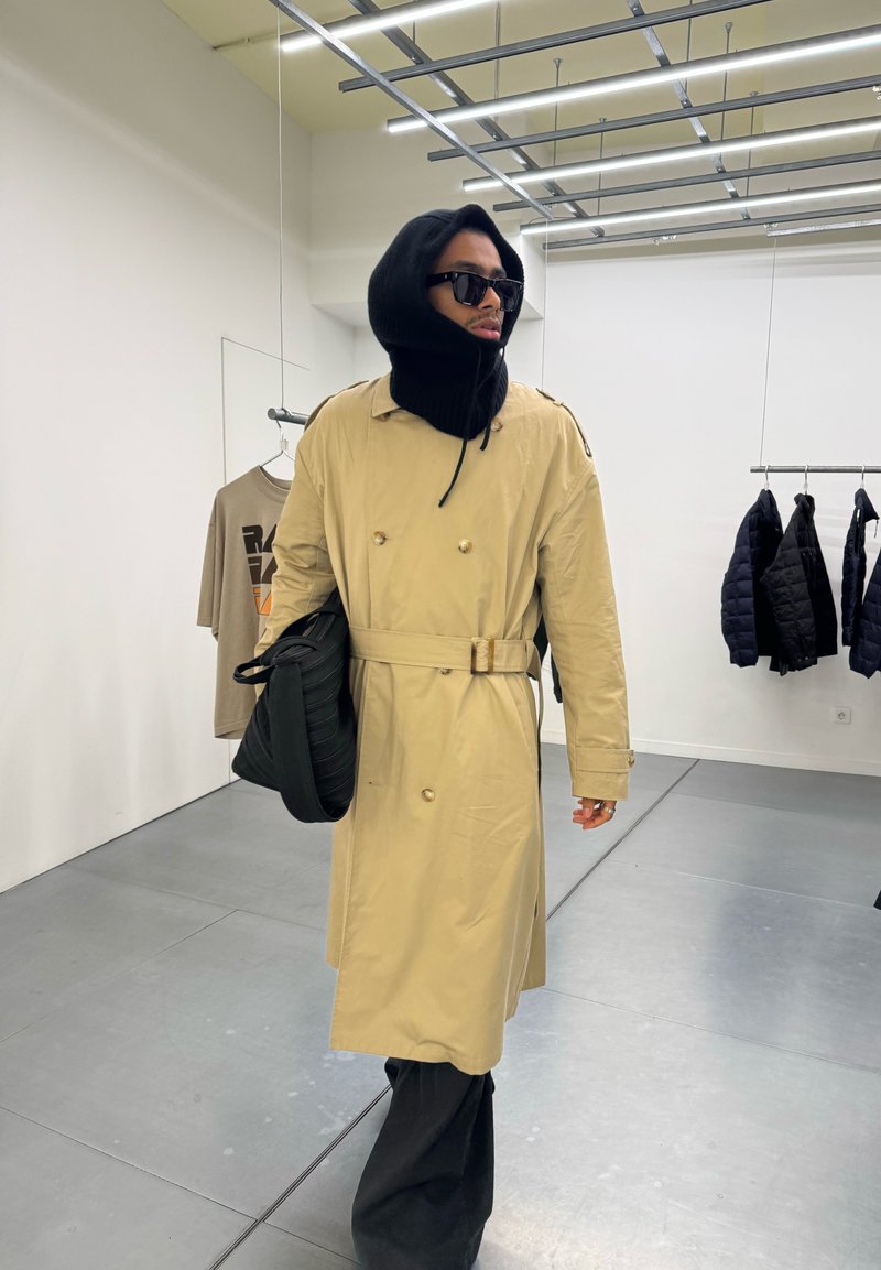 Person iført beige trenchcoat, svart hette, solbriller og holder en svart veske inne i en klesbutikk med jakker og skjorter utstilt.