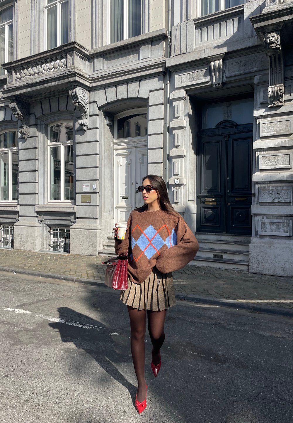 Een bruine trui met een blauw en rood ruitpatroon, een geplooide beige rok, rode puntige hakken en een rode tas, terwijl ze een kop koffie vasthoudt.