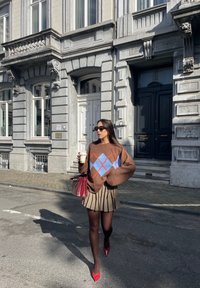 Een bruine trui met een blauw en rood ruitpatroon, een geplooide beige rok, rode puntige hakken en een rode tas, terwijl ze een kop koffie vasthoudt.