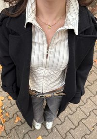 Fekete kabát, fehér és szürke csíkos cipzáras ing, szürke nadrág, ezüst tornacipő, arany szív alakú nyaklánc. Texturált szövet részletek láthatók.