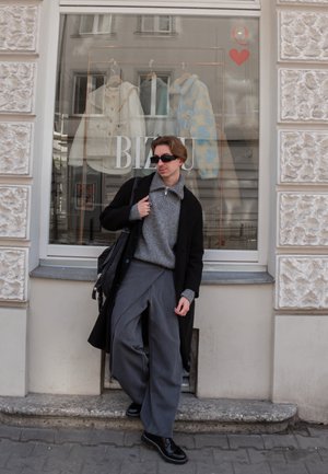 Joven con abrigo negro, suéter gris, pantalones anchos y gafas de sol apoyado contra el escaparate de una tienda con abrigos colgados.