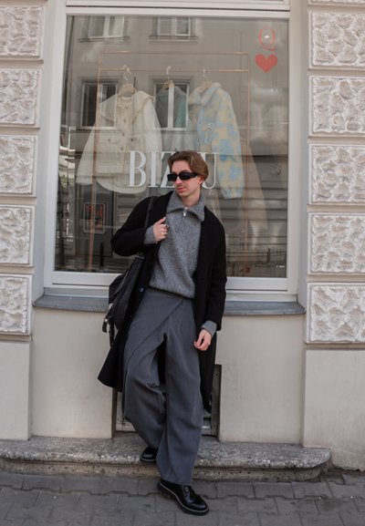 Joven con abrigo negro, suéter gris, pantalones anchos y gafas de sol apoyado contra el escaparate de una tienda con abrigos colgados.