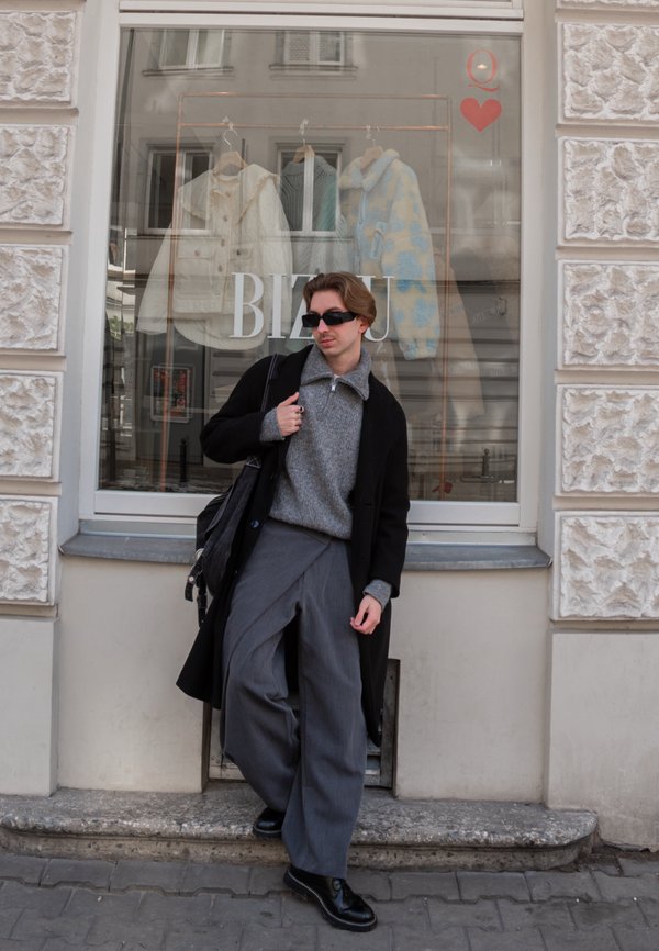 Mladá osoba oblečená v čiernom kabáte, sivom svetri, širokých nohaviciach a slnečných okuliaroch sa opiera o výkladnú skriňu s visiacími kabátmi.