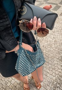 Mazs tirkīza krāsas rokassomu ar sudraba uzlīmēm, melni saulesbrilles ar zelta akcentiem un melna Ray-Ban somiņa, kas turēta rokā ar krāsotiem nagiem.