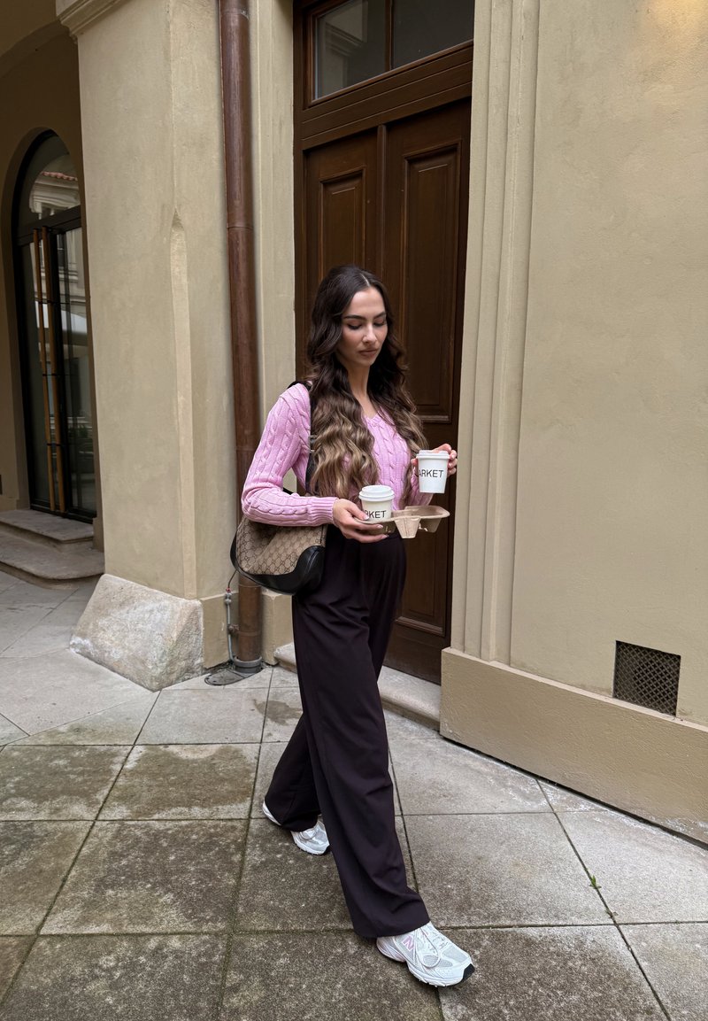 Femme en pull rose et pantalon noir marche sur un pavé en pierre en portant deux tasses de café dans un porte-gobelets près de l'entrée d'un bâtiment beige.