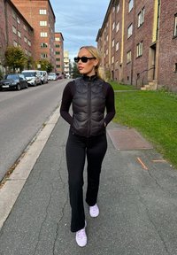 Μαύρο γιλέκο puffer με φερμουάρ και κατακόρυφη ραφή, συνδυασμένο με μαύρες φαρδιές παντελόνες και ανοιχτό μωβ αθλητικά παπούτσια, περπατώντας σε έναν δρόμο.