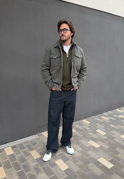 Hombre con gafas que lleva una chaqueta verde, un forro polar oliva, pantalones vaqueros anchos oscuros y zapatillas blancas, de pie sobre un pavimento de baldosas junto a una pared gris.