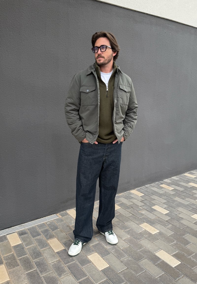 Man with glasses wearing green jacket, olive fleece, dark wide-leg jeans, and white trainers standing on tiled pavement by grey wall.