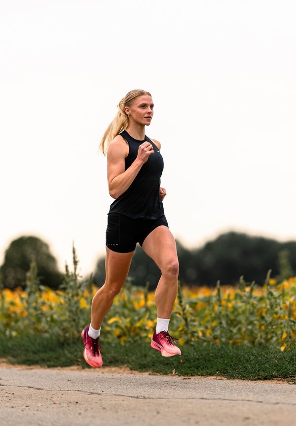 Femme en tenue de sport noire et chaussures roses courant à l'extérieur sur un chemin avec des plantes vertes et des fleurs jaunes en arrière-plan.