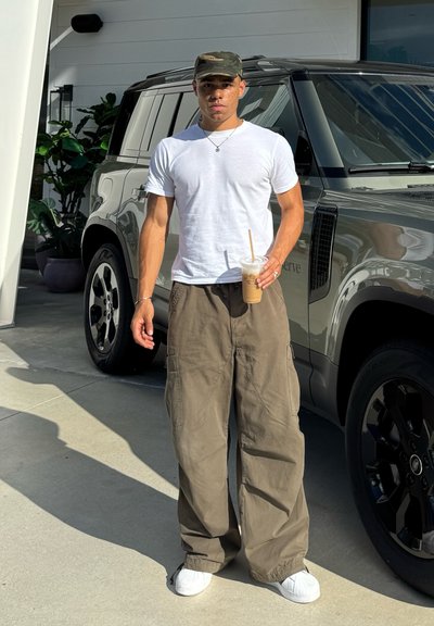 Camiseta blanca, pantalones cargo verde oliva y zapatillas blancas. Sosteniendo una bebida para llevar. Vehículo gris aparcado de fondo.