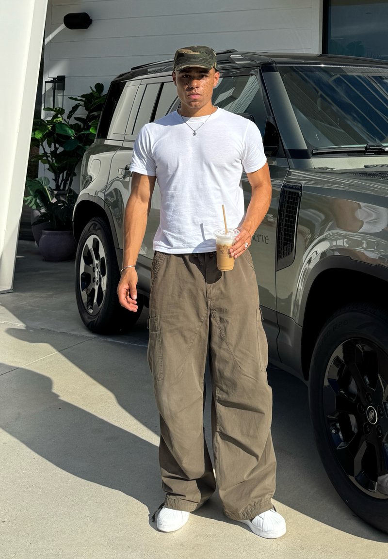 Maglietta bianca, pantaloni cargo verde oliva e sneakers bianchi. Tiene una bevanda da asporto. Veicolo grigio parcheggiato sullo sfondo.