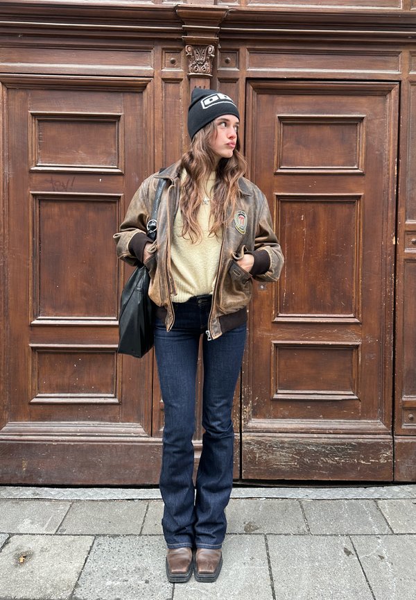 Ung kvinne i brun skinnjakke, beige genser, mørke jeans og svart lue står med hendene i lommene foran store tredører.