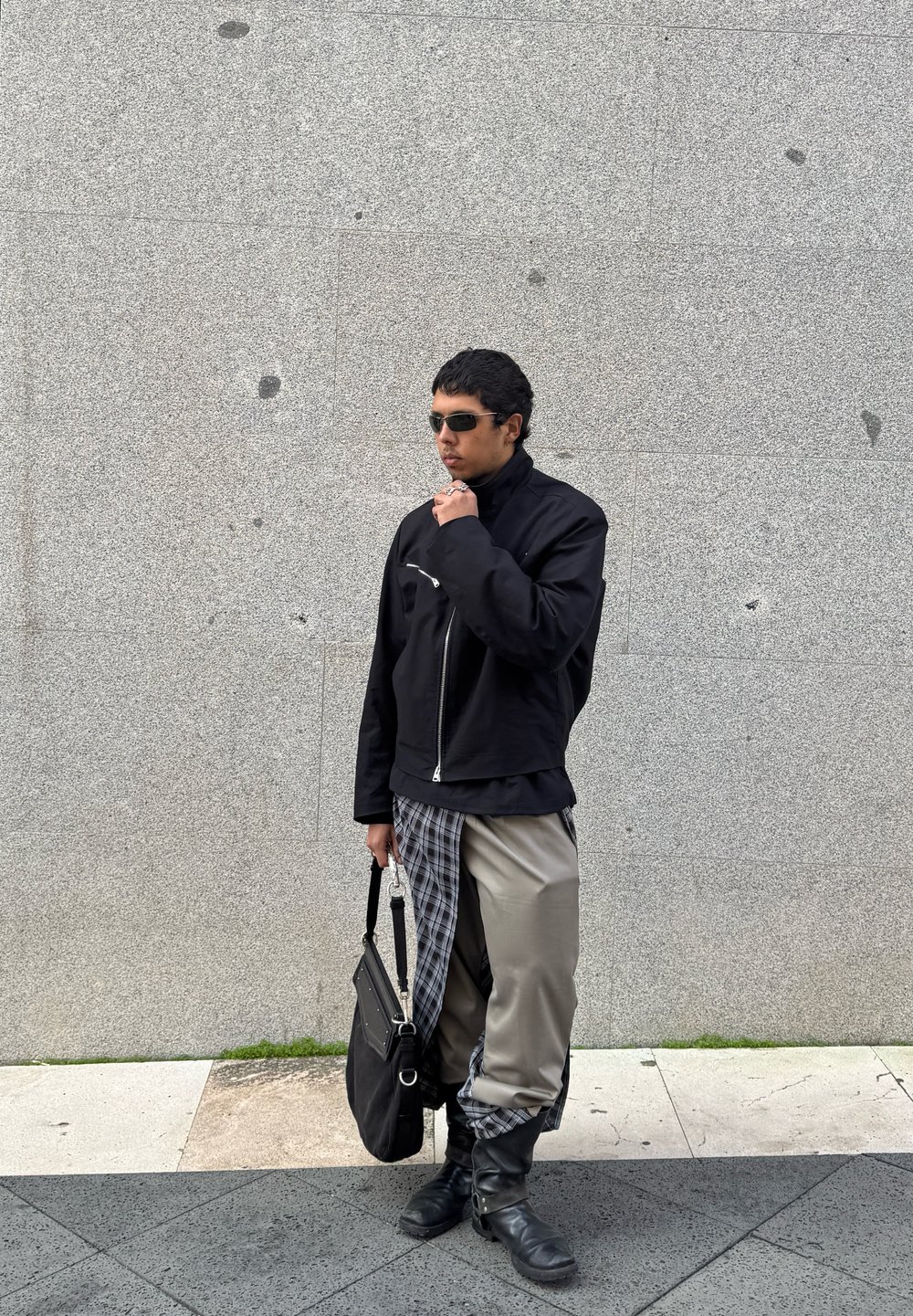 Μαύρο τζάκετ με φερμουάρ και ψηλό γιακά, γκρι καρό παντελόνι και μαύρες τρύπιες μπότες. Κρατά μια μαύρη τσάντα ώμου. Λείος υφ�ασμάτινος, μοντέρνος σχεδιασμός.