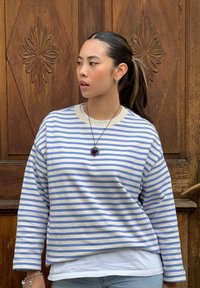 Ριγωτό πουλόβερ σε λευκό και ανοιχτό μπλε, με χαλαρή εφαρμογή, στρογγυλή λαιμόκοψη και μακριά μανίκια, φορεμένο πάνω από λευκό πουκάμισο.