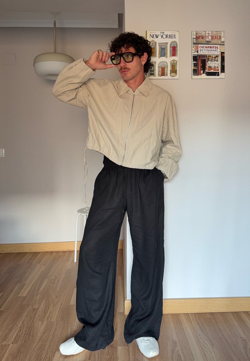 Homme aux cheveux bouclés et moustachu ajustant de grandes lunettes de soleil noires, portant une veste beige zippée, un pantalon large noir et des chaussures blanches à l'intérieur.
