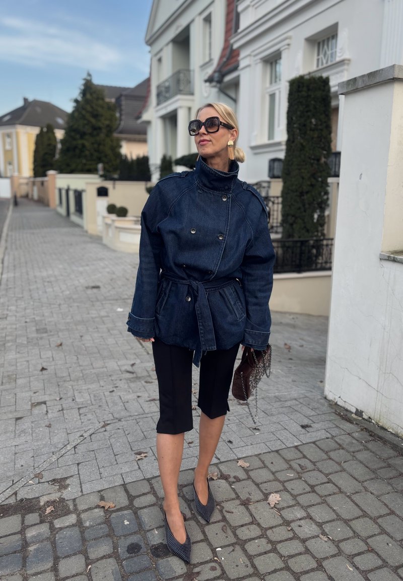 Femeie blondă purtând o geacă supradimensionată din denim închis, pantaloni negri scurți, pantofi cu vârf ascuțit, ochelari de soare mari, ținând o geantă maro pe o stradă pavată.