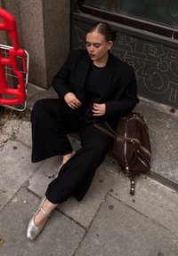 Black oversized blazer worn over a black top, paired with wide-leg black trousers, silver strap shoes, and a brown suede bag, positioned on a stone pavement. A red barrier is visible in the background.