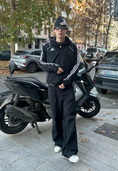Chaqueta deportiva negra de Adidas con rayas blancas, pantalones anchos negros y zapatillas blancas. Accesorios incluyen gafas de sol y un gorro. Motocicleta aparcada cerca.