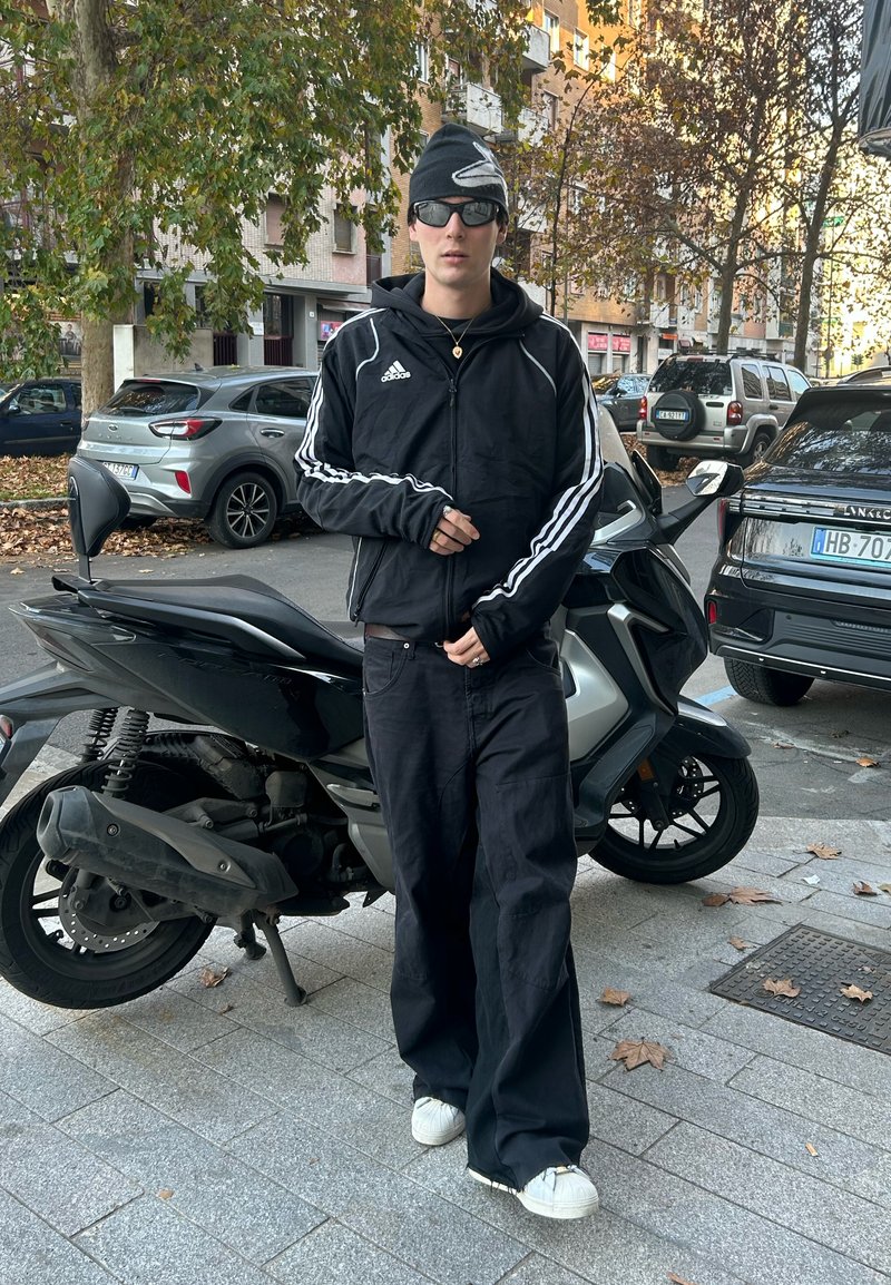 Sort Adidas træningsjakke med hvide striber, sorte brede bukser og hvide sneakers. Tilbehør inkluderer solbriller og en beanie. Parkeret motorcykel i nærheden.